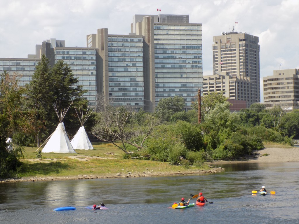 A true contrast in what the Ottawa River means to different people.  Young kayakers enjoy a hot afternoon by Victoria Island where tepees and a totem pole identify it as a sacred place for the Algonquin.  Government office buildings in Gatineau, Quebec loom in the background.  Photo by James Morgan